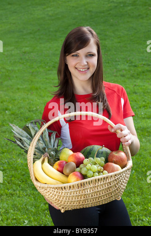 Jeune femme portant des vêtements de sport, souriant et tenant un panier de fruits Banque D'Images