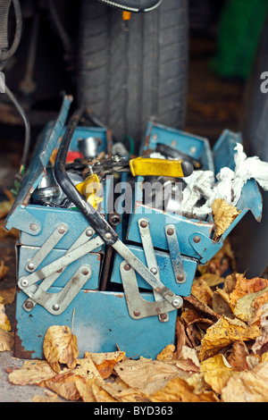 Une vieille boîte à outils en métal rouillé bleu se trouvant sur le plancher d'un garage, parmi un tas de feuilles mortes, et en face d'un pneu de voiture. Banque D'Images