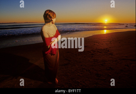 Femme sur la plage au coucher du soleil enveloppée dans un châle. Banque D'Images