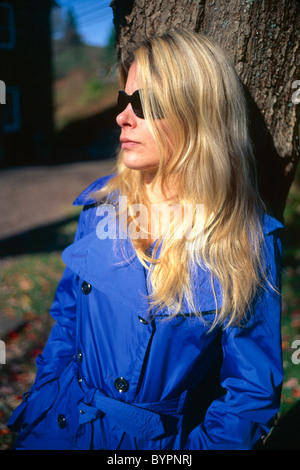 Jeune femme blonde en manteau bleu portant des lunettes de soleil, appuyé contre un arbre, Automne, New Jersey Banque D'Images