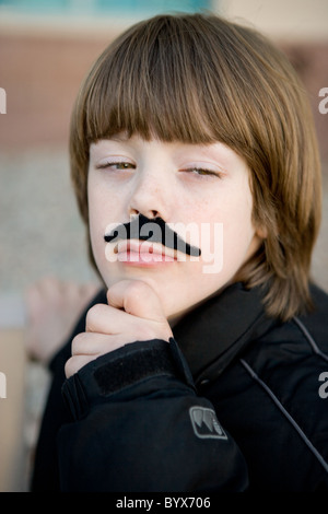 Un garçon de huit ans portant un semblant de moustache with hand on chin, agissant comme un homme adulte dans une profonde réflexion. Banque D'Images