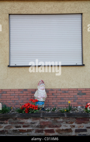 Weathered garden gnome devant une fenêtre avec des stores fermés Banque D'Images