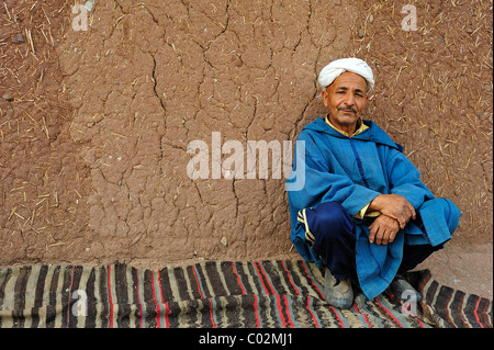 Portrait d'un homme âgé portant une Djellabah traditionnelle et un turban assis sur une couverture et appuyé contre le mur de boue Banque D'Images