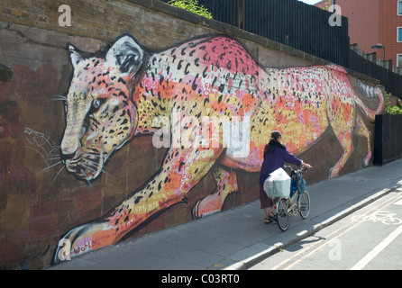 Une femme poussant son vélo d'une route, le long d'un mur peint avec une rose et jaune leopard, à Londres Banque D'Images