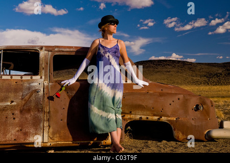 Femme portant des gants de bal blanc, Tye est mort, robe et holding Red Rose, s'appuie contre la voiture criblée de balles oid rouillées. Banque D'Images