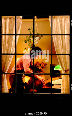 Nuit vue à travers une fenêtre d'une femme avec son Golden Retriever dog travaillant sur un ordinateur à la maison Banque D'Images