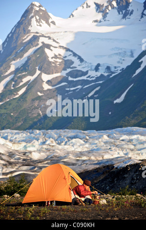 Femme prépare la nourriture sur une cuisinière de randonnée alors que dans sa tente, Spencer Glacier en arrière-plan, la Forêt Nationale de Chugach, Alaska Banque D'Images