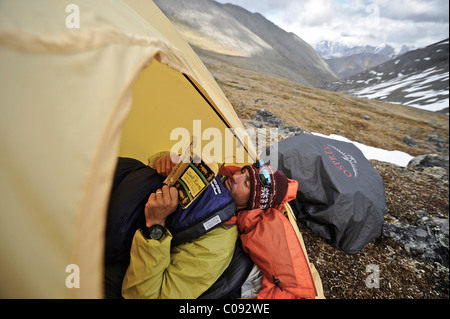 Backpacker lit un livre à l'intérieur d'une tente et attend le mauvais temps à un camp alpin Mt ci-dessous. Chamberln, Brooks, RFNA Banque D'Images