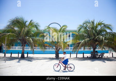 Une petite fille rides son vélo sur le sable en face d'un bâtiment coloré et d'une rangée de palmiers. Banque D'Images