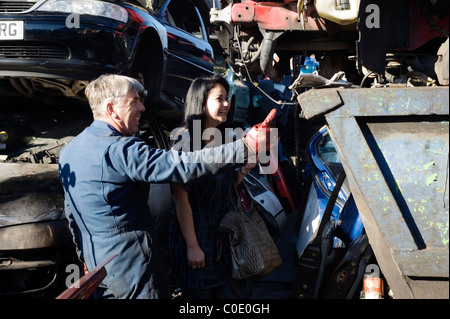 Travailleur ferraille montrant jeune femme pièce de rechange sur voiture ferraille Banque D'Images