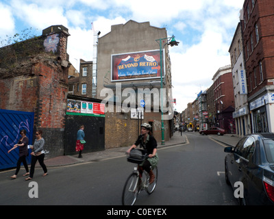 Au-delà du panneau d'affichage rétro affiche et femme avec un casque à vélo et piétons un dimanche sur un Brick Lane East End vide à Londres Angleterre Royaume-Uni KATHY DEWITT Banque D'Images