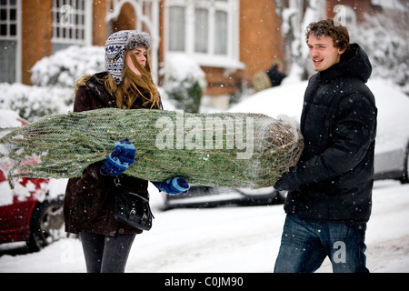 Un jeune couple transportant un arbre de Noël le long d'une rue enneigée Banque D'Images