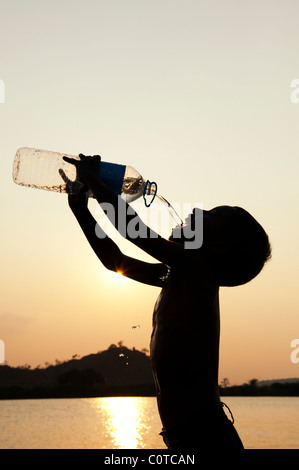 Jeune Indien boire de l'eau minérale à partir de la bouteille d'eau en plastique au coucher du soleil. Silhouette. L'Inde Banque D'Images