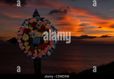 Un mémorial en bord de route avec une couronne florale sur une croix rend hommage à une victime d'accident de voiture, silhouettée contre un coucher de soleil éclatant sur l'océan, les açores. Banque D'Images