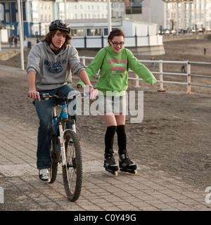 Un adolescent à cheval sur un vélo et une fille faire du roller sur la promenade de aberystwyth wales uk Banque D'Images