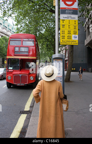 Une femme dans un long manteau brun et blanc hat attend un bus routemaster rouge à l'arrêt sur le brin dans le centre de Londres, au Royaume-Uni. Banque D'Images