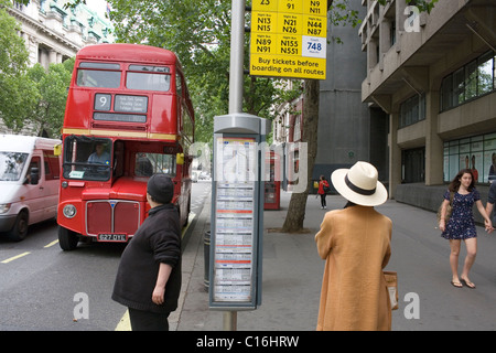 Une femme dans un long manteau brun et blanc hat attend un bus routemaster rouge à l'arrêt sur le brin dans le centre de Londres, au Royaume-Uni. Banque D'Images