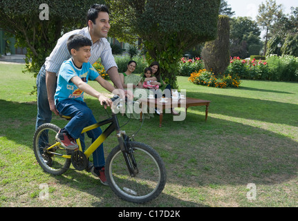 L'enseignement de l'homme son fils pour circuler à bicyclette avec sa famille assis dans l'arrière-plan Banque D'Images