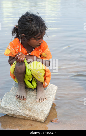 Young mauvaise caste inférieure Indian street girl accroupis sur un rocher dans une rivière. L'Andhra Pradesh, Inde Banque D'Images