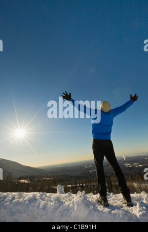 Un male hiker avec bras tendus sur une crête surplombant Anchorage, Alaska avec soleil et montagnes en arrière-plan. Banque D'Images