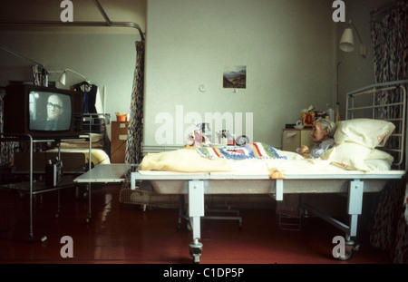 Une femme âgée patiente avec un châle crocheté assis dans un lit regardant la télévision dans un hôpital NHS dans les années 1980 Royaume-Uni Grande-Bretagne KATHY DEWITT Banque D'Images