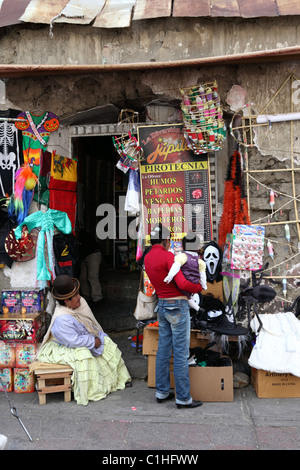 Aymara femme assise à l'extérieur de l'entrée du magasin vendant des masques de feux d'artifice et des costumes pour Halloween, la Paz, Bolivie Banque D'Images