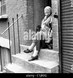 1960, Londres, une vieille dame au foulard portant un imperméable ou mac assis sur une pierre l'étape d'une maison victorienne avec terrasse. Banque D'Images