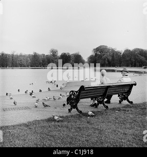 Années 1950, tôt le matin et un couple de personnes âgées assis sur un banc à côté du lac de navigation vide au Royal Park, Kensington Gardens, Londres, Angleterre, Royaume-Uni. Banque D'Images