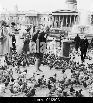 Années 1960, Trafalgar Square, Londres. Les pigeons entourent une visiteur de dame, tandis qu'un jeune garçon obtient une boîte de pois secs d'un stalle pour nourrir les oiseaux. Banque D'Images