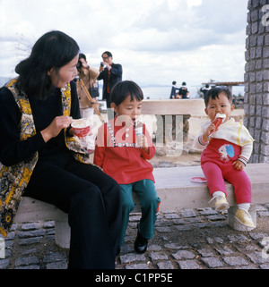 Photo de 1970 par J Allan Paiement d'une femme japonaise avec deux jeunes enfants assis sur le banc en béton de faible, de manger des glaces. Banque D'Images