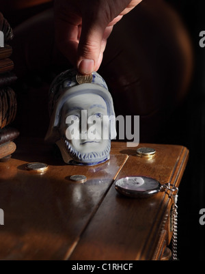 Ancienne banque en porcelaine hollandaise en forme de tête d'homme avec pièces de monnaie et montre de poche vintage sur la table et la main pièce de monnaie en banque Banque D'Images