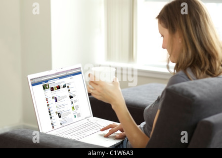Femme se prélasser dans le salon lors de la vérification de son compte Facebook sur un ordinateur portable Banque D'Images