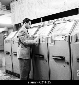 1950s, un homme en costume et cravate, un homme nouvellement arrivé immigré en Grande-Bretagne, utilisant une machine à billets à pièces dans le métro de Londres, Angleterre, Royaume-Uni. Banque D'Images