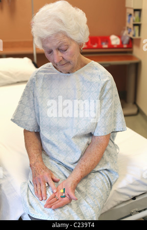 Une vieille dame dans une chemise d'hôpital:assis sur un lit:en regardant plusieurs pilules dans sa main Banque D'Images