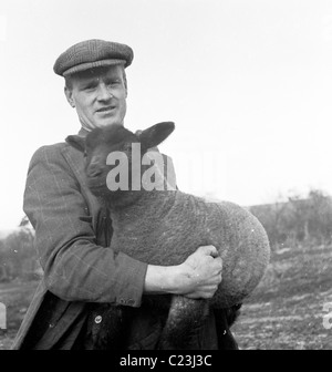 Années 1950, l'Angleterre. Un agriculteur détient un mouton noir dans ses bras dans ce tableau historique par J Allan l'argent comptant. Banque D'Images