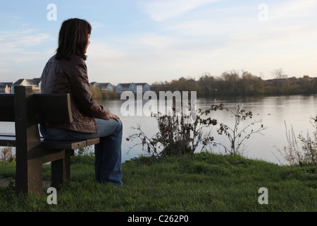 Vue arrière d'une femme profondément dans ses pensées à la recherche vers un lac, assis sur un banc de parc. Banque D'Images