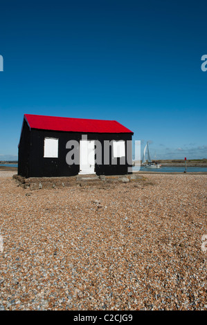 Un yacht blanc voiles passé une cabane en carton ondulé rouge et noir avec portes blanches construit sur la plage de galets à Rye Harbour. East Sussex Banque D'Images