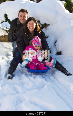 Portrait vertical d'une jeune famille, maman, papa et de deux jeunes filles qui jouent tous ensemble sur une luge ou un traîneau dans la neige Banque D'Images