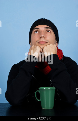 Jeune homme pensif dans les vêtements d'hiver assis à table et jusqu'à la sur fond bleu Banque D'Images