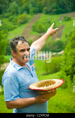 Farmer holding son chapeau de paille et montrant le labour des terres sur la campagne collines à un espace vert avec les terres cultivées et les forêts de nombreux Banque D'Images