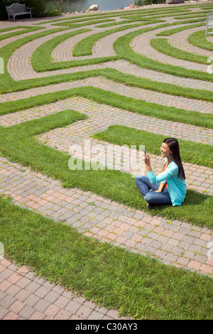 Belle jeune femme asiatique rit d'un message texte sur son portable tout en restant assis dans un labyrinthe d'herbe. Banque D'Images