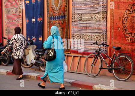 Les femmes voilées en passant devant des tapis berbères, l'ENTRÉE À L'Bazar (tapis) DU MARCHÉ EN PLEIN CŒUR DE LA MÉDINA, Marrakech, Maroc Banque D'Images