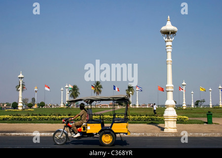 Un homme conduisant un tuk tuk est un groupe de drapeaux qui passe près de la promenade Riverside sur le Mékong à Phnom Penh, Cambodge. Banque D'Images