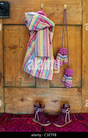 Les jeunes filles manteau et mitaines à rayures colorées suspendu par une cuisine en bois avec porte éraflée bottes sur le mat Banque D'Images