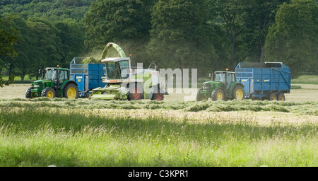 2 tracteurs John Deere et remorques de l'Ouest travaillant et conduisant dans les champs agricoles avec ensileuse Claas, chargement d'herbe coupée (ensilage) - Yorkshire, Angleterre, Royaume-Uni. Banque D'Images