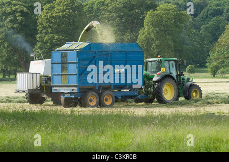 1 tracteur et remorque John Deere travaillant dans les champs agricoles, roulant le long de l'ensileuse Claas récoltant de l'herbe coupée (ensilage) - Yorkshire, Angleterre, Royaume-Uni. Banque D'Images