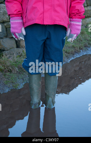 Jeune enfant (fille) en bottes wellington vertes (bottes en caoutchouc) imperméable rose, gants et pantalon bleu debout dans une flaque boueuse profonde (jambes et corps) - Angleterre. Banque D'Images