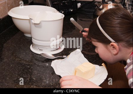 Une jeune fille qui cuit (tête et mains) pèse du beurre sur des balances, se plie pour lire le poids. Couteau à la main, cuillère, verseuse et bol (ustensiles de cuisine) - Angleterre Royaume-Uni. Banque D'Images