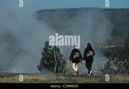 Deux touristes avec USA drapeau sur leur dos dans le parc de Yellowstone geyser en éruption de vapeur avec, Upper Geyser Basin, Old Faithful, le parc de Yellowstone, États-Unis Banque D'Images