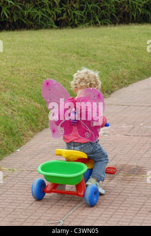 Blondie petite fille aux cheveux bouclés courts portant des ailes de papillon debout avec tricycle sur l'herbe dans un parc. Banque D'Images